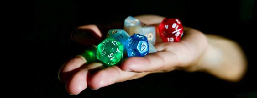 Reaching out from the shadows, a hand holds a collection of multicolored, polyhedral dice.